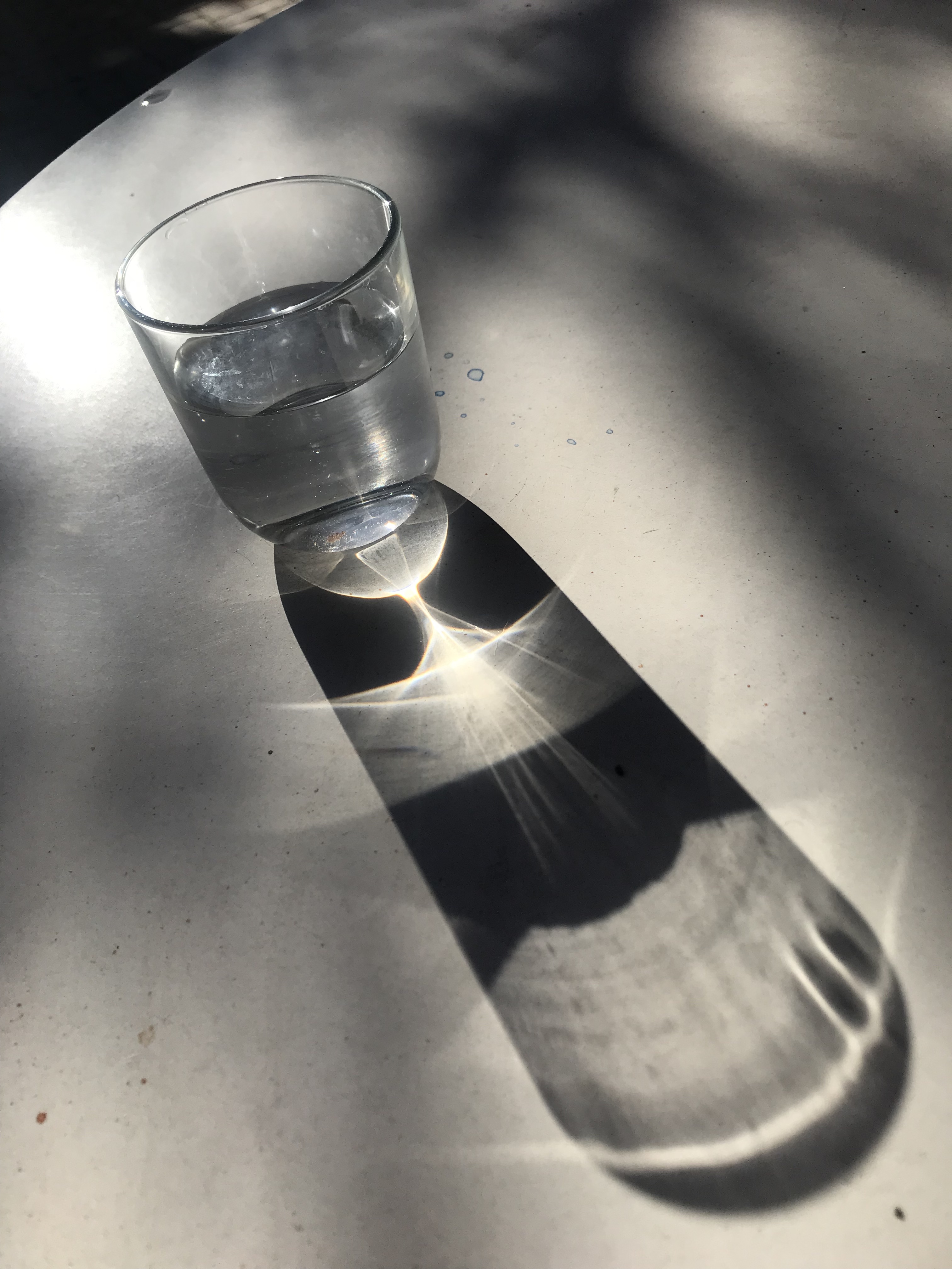 A morning photo of a glass of water, with low solar angle sunglight illuminating the glass, and water, creating awesome elongated shadows and mathematical patterns. Shadows of a tree dance on the table, and the colors mix. 